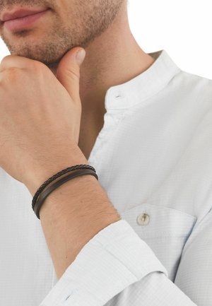 Hombre con camisa blanca de cuello mandarín tocándose la barbilla, con la muñeca mostrando dos pulseras de cuero marrón oscuro.