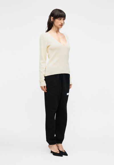 Ανοιχτό μπεζ πουλόβερ με V λαιμόκοψη, μαύρο φαρδύ παντελόνι με ελαστικές μανσέτες και μαύρες γόβες με μυτερή μύτη. Λείο ύφασμα, μινιμαλιστικός σχεδιασμός.
