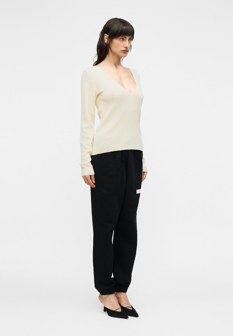 Pull beige clair à col en V, pantalon noir ample avec poignets élastiques, et escarpins noirs à bout pointu. Texture lisse, design minimaliste.