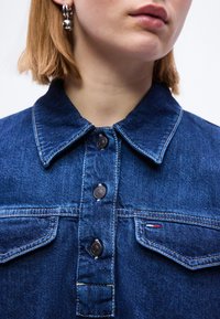 Κοντινό πλάνο ενός ατόμου που φοράει μπλε τζιν μπουφάν με μεταλλικά κουμπιά και ασημένια σκουλαρίκια κρίκους, εμφανίζοντας μέρος του λαιμού και των χειλιών.