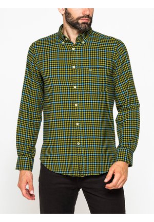 Uomo che indossa una camicia a maniche lunghe abbottonata con motivo a quadri verde, nero e blu e pantaloni neri, in piedi davanti a uno sfondo bianco.