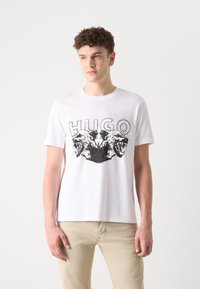 Hvit bomulls T-skjorte med et dristig svart grafisk trykk av to stiliserte dyrehoder og teksten "HUGO" over dem.