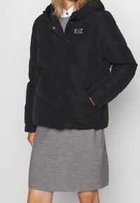 Chaqueta de plumas negra con capucha, con un patrón de rombos acolchados, cierre de cremallera y logo de EA7. La tela parece suave y mate.