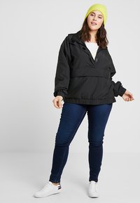 Juodas lengvas džemperis su pusiau užtrauktuku, elastinėmis rankovėmis ir susitraukusiu kraštu, dėvimas ant baltos marškinėlių, derinamas su tamsiomis džinsomis ir baltomis sportbačiais.