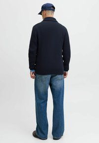 Marine gebreide cardigan met een sjaalkraag en geribbelde zoom, gecombineerd met lichtblauwe wijde jeans. Een donkerblauwe pet maakt de outfit compleet.