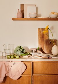 Blat kuchenny z różowym fartuchem w kształcie kota, świeżymi warzywami, szklanym dzbankiem, metalowymi pojemnikami, drewnianymi deskami do krojenia i elementami dekoracyjnymi.