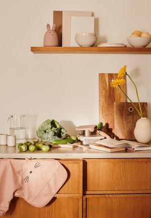 Blat de bucătărie cu un șorț roz în formă de pisică, legume proaspete, carafă din sticlă, recipiente metalice, tocătoare din lemn și obiecte decorative.