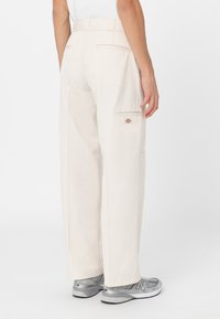 Pantalones de pierna ancha beige, fabricados en un material resistente, con dos bolsillos traseros y un pequeño logo en el lateral. Textura suave con un corte recto.