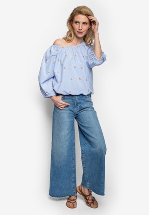 Blonde Frau trägt eine schulterfreie hellblaue Bluse mit orangefarbenen Blumenmustern, weite Jeans und braune Sandalen, posiert mit der Hand im Haar.