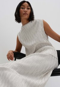 Mouwloze, geplooide jurk in lichtbeige, met een hoge halslijn en een vloeiende snit, gecombineerd met een zwarte stoel.