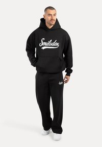 Sudadera negra con el logo blanco "Smilodx", bolsillo canguro delantero, pantalones de chándal negros a juego y zapatillas blancas. Tela suave y ajuste holgado.