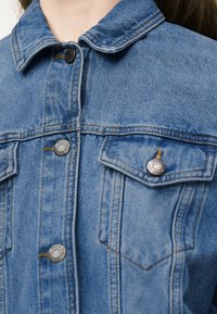 Nærbillede af en blå denimjakke med metalknapper og brystlommer, båret af en person.