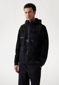 Chaleco negro con cremallera y capucha, que cuenta con múltiples bolsillos, un sistema de correas en un hombro y un parche con logo. Textura de tejido técnico y suave.
