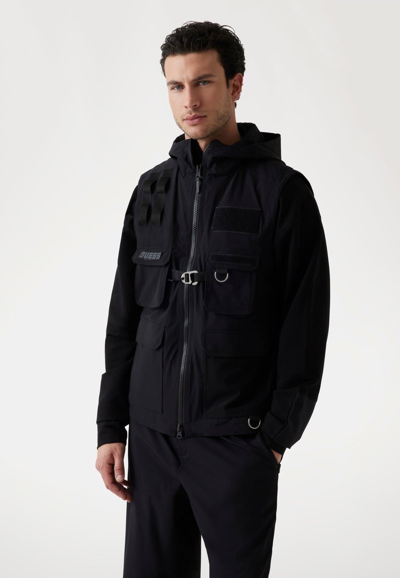 Chaleco negro con cremallera y capucha, que cuenta con múltiples bolsillos, un sistema de correas en un hombro y un parche con logo. Textura de tejido técnico y suave.