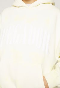 Sudadera amarilla clara con una textura suave, que presenta una gran impresión blanca de "PEGADOR" en la parte frontal y un bolsillo tipo canguro.