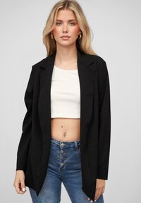 Fekete blézer, kivágott gallérral, hosszú ujjakkal és nyitott elöl, fehér crop top és magas derekú kék farmer nadrág felett viselve.