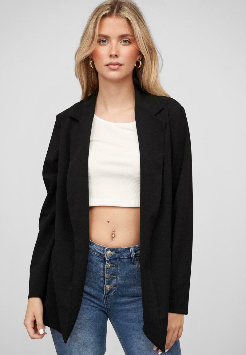 Fekete blézer, kivágott gallérral, hosszú ujjakkal és nyitott elöl, fehér crop top és magas derekú kék farmer nadrág felett viselve.