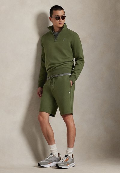 Φούτερ με φερμουάρ στο χρώμα του ελαιόχρωμου με ριμπ μανσέτες, ταιριαστά σορτς και γκρι αθλητικά παπούτσια με λευκές σόλες. Στυλιζαρισμένο με σκούρα γυαλιά ηλίου.