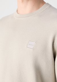 Pull beige col rond avec un petit logo carré « BOSS » sur la partie gauche de la poitrine.