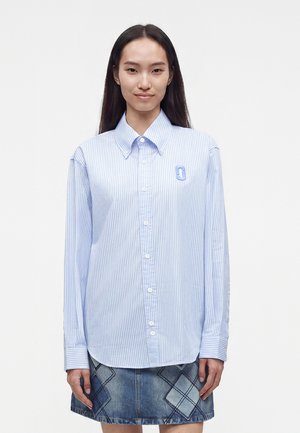 Femme debout et faisant face, vêtue d'une chemise bleue à rayures boutonnée avec un logo et d'une jupe en jean bleue à motifs losanges.