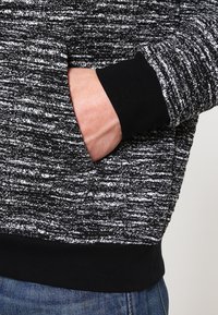 Svart och vit texturerad sweatshirt med långa ärmar, med en ficka och ribbad svart mudd. Tyget har ett vävt, prickigt mönster.