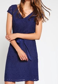 Námořnická modrá krajková šaty s krátkými rukávy, véčkovým výstřihem a saténovým páskem v oblasti pasu. Obsahují texturovaný květinový vzor a délku po kolena.