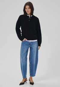 Frau trägt einen schwarzen Polo-Strickpullover mit Knöpfen, hellblaue Jeans mit hoher Taille und schwarze Slingback-Pumps mit niedrigem Absatz, steht vor einem einfarbigen Hintergrund.