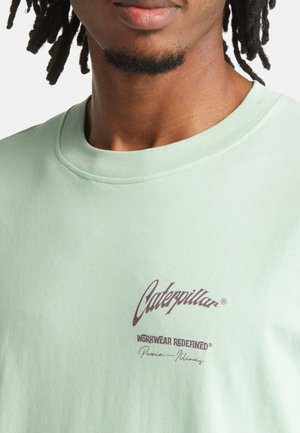 Muntgroen katoenen T-shirt met een ronde halslijn en een bedrukt logo in bordeauxrood met de tekst "Caterpillar Workwear Redefined."