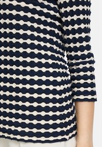 Marineblau und weiß gestreiftes Oberteil mit einem strukturierten, welligen Muster, das über drei Viertel Ärmel und ein tailliertes Design verfügt.