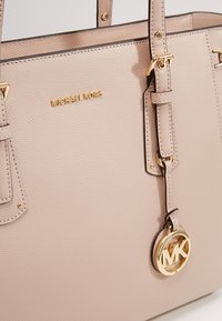 Sac à main en cuir beige Michael Kors avec des lettres dorées du logo et un charm circulaire MK suspendu à une bandoulière.