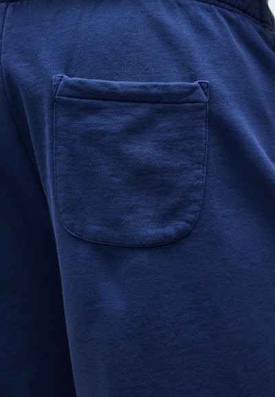 Κοντινή λήψη μιας τσέπης από ύφασμα σκούρου μπλε ραμμένης σε ρούχο του ίδιου χρώμα τος, με έμφαση στην υφή και τις λεπτομέρειες των ραφών.