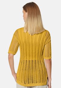 Gelbes Strickpullover mit kurzen Ärmeln, charakterisiert durch ein offenes Häkelmuster und einen runden Saum, strukturierte Oberfläche und lockere Passform.