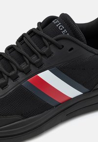 Chaussure de sport noire avec empeigne en mesh texturé et semelle en caoutchouc, agrémentée de bandes rouges, blanches et bleues sur le côté.