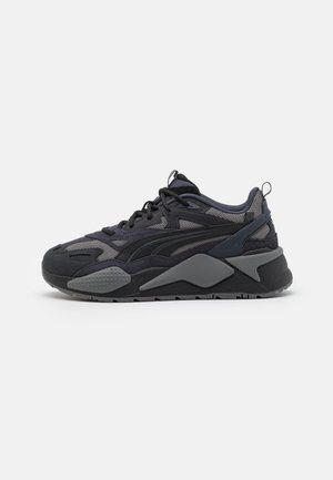 Zapatilla deportiva negra y gris de corte bajo con paneles de malla y ante, suela gruesa, lengüetas para tirar y cordones negros, mostrada de lado sobre fondo blanco.