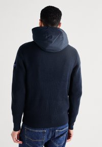 Granatowy sweter robiony na drutach z odpinanym, pikowanym kapturem, prążkowaną fakturą i dopasowanym krojem. Podkreślony naszywką z logo na lewym ramieniu.