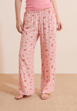 Vrouw draagt losse roze pyjamabroek met kleine rode kersenpatronen, staat blootsvoets op een beige tapijt.