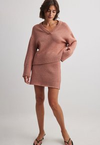 Roza pletena jopica z V-izrezom in rebrastim robom, v kombinaciji z usklajenim pletenim mini krilom. Mehka tekstura z diskretnimi sijočimi poudarki.