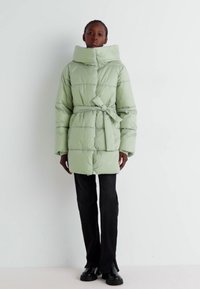 Manteau matelassé vert clair avec un col montant, une taille cintrée par une ceinture, un motif matelassé et des poches latérales, assorti à un pantalon noir et des bottes.