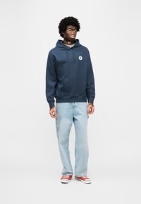 Μπλε φούτερ με κουκούλα, τσέπη καγκουρό και λογότυπο, συνδυασμένο με ανοιχτόχρωμο τζιν και κόκκινα αθλητικά παπούτσια. Ομαλή υφή και άνετη εφαρμογή.