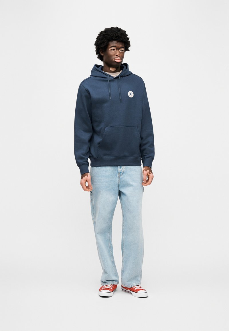 Marineblå hættetrøje med kængurulomme og logopatch, kombineret med lyseblå jeans og røde sneakers. Glat tekstur og afslappet pasform.