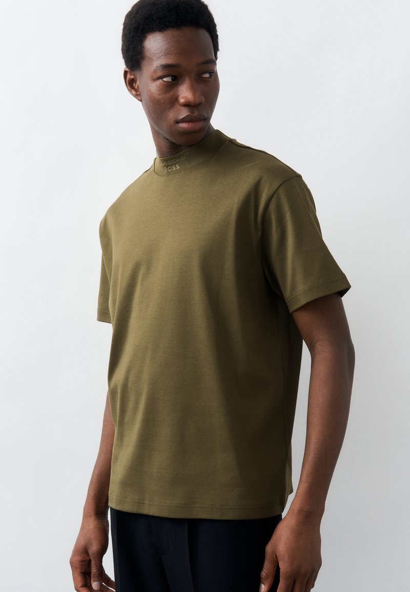 Νεαρός άνδρας που φοράει ελιά πράσινο κοντομάνικο πουκάμισο με ένα μικρό λογότυπο "BOSS" στον γιακά, κοιτάζοντας προς τα αριστερά του με φόντο μια απλή επιφάνεια.