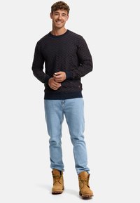Pull en bleu marine à motifs avec un design en losanges, manches côtelées et col rond, associé à un jean bleu clair et des bottes beiges.