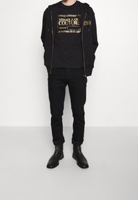 Sudadera negra con cremallera y acentos dorados, camiseta negra con texto dorado, jeans negros y botas de cuero negras. Conjunto simple y monocromático.