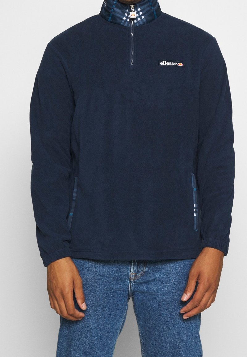 Pull-over en polaire bleu marine avec une fermeture éclair au quart, des poignets élastiques et des poches latérales. Présente un col à motif à carreaux et un logo sur la poitrine.