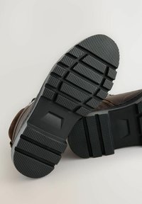 Braune Wildlederstiefel mit einer schwarzen Gummisohle, die ein strukturiertes, gemustertes Profil für besseren Halt aufweist. Seitlicher Reißverschluss an der Oberseite.