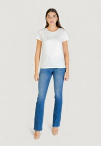 Biała koszulka z napisem „LIU·JO” wykonanym z kryształków, zestawiona z niebieskimi dzwonami. Modelka nosi beżowe szpilki, prezentując prosty i swobodny strój.