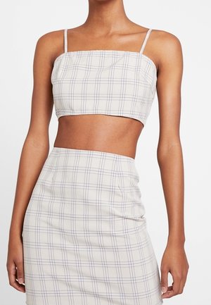 Vrouw draagt een wit geruit crop top met dunne bandjes en een bijpassende rok met hoge taille, waarbij de buik zichtbaar is tegen een effen achtergrond.