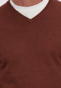 Persoon in een bruin V-hals sweater over een witte ronde hals t-shirt, zichtbaar zijn alleen het bovenste deel van de borst en de hals.