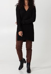 Czarny sweter z prążkowanej dzianiny z dekoltem w serek, z bocznymi rozcięciami, w połączeniu z brązowymi dopasowanymi spodniami z zamszu i czarnymi botkami na obcasie. 