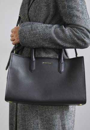 Persoon in een grijze visgraatjas houdt een zwarte gestructureerde leren handtas van Michael Kors met twee handvatten vast.